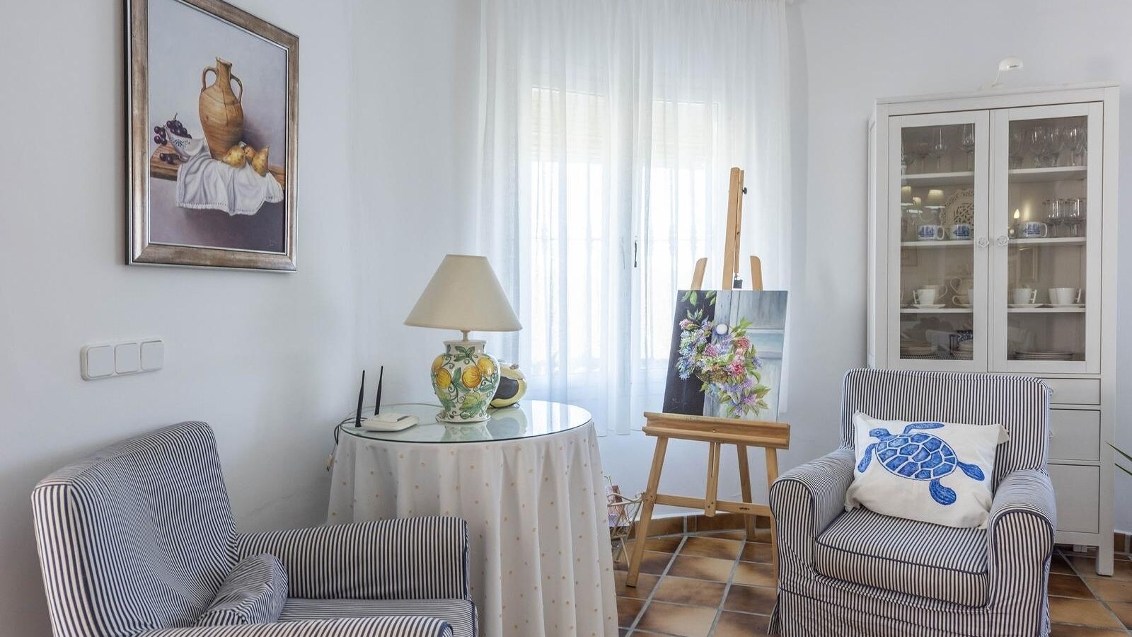 A room with striped chairs, a table with a lamp, a painting, and a cabinet with glass doors.