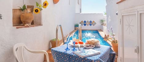 A small outdoor dining area with a table set for two, a pool, and potted plants.