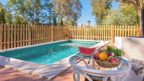 A swimming pool with a wooden fence, a table with fruits and a watermelon, and a lamp post in the background.