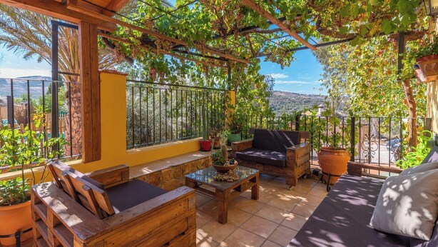 A sunlit patio with wooden furniture, a stone bench, and potted plants.