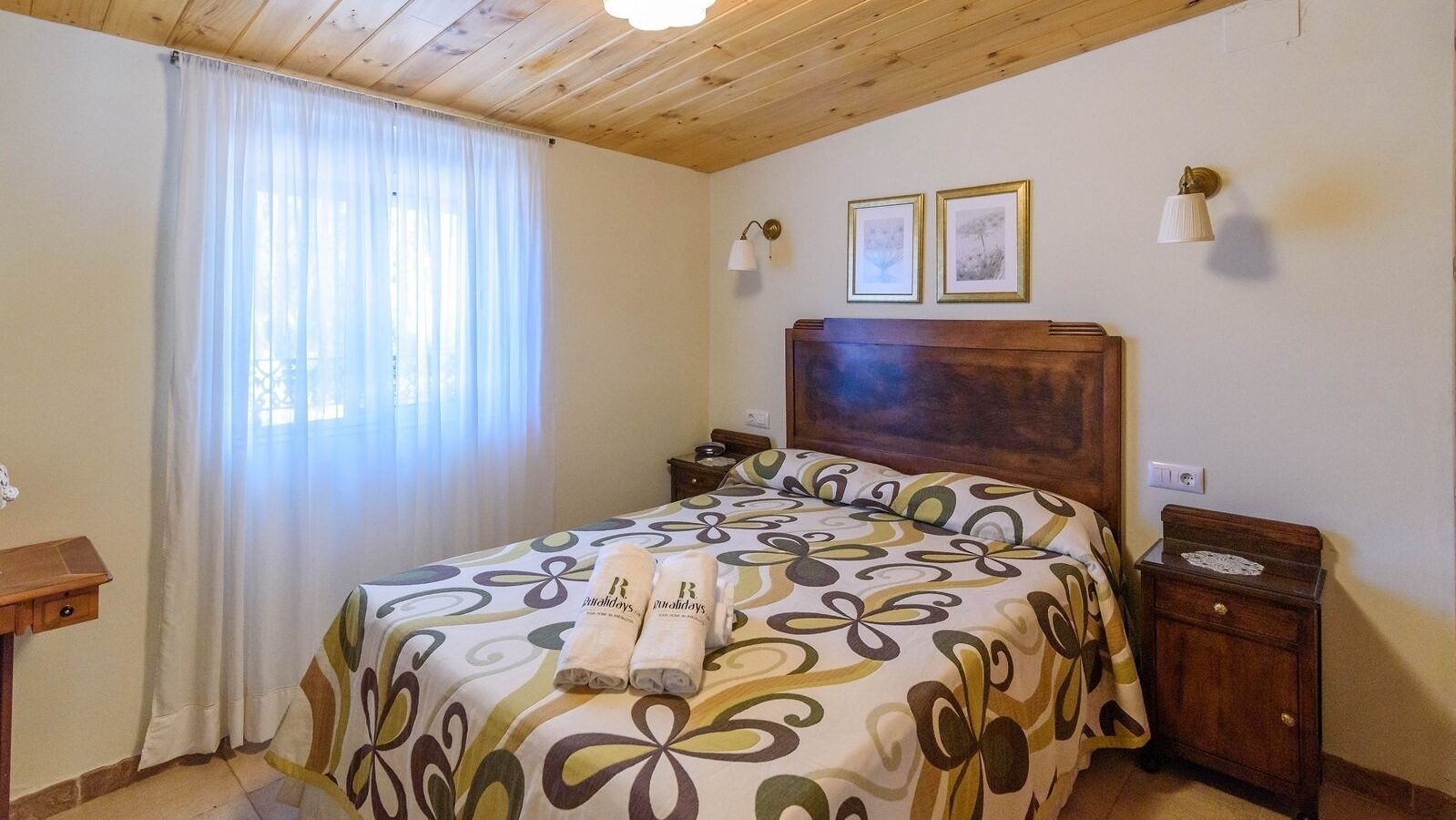 Un dormitorio con techo de madera, una cama con colcha floral, dos cuadros en la pared y una mesita de noche con una lámpara.