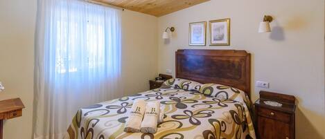 Un dormitorio con techo de madera, una cama con colcha floral, dos cuadros en la pared y una mesita de noche con una lámpara.
