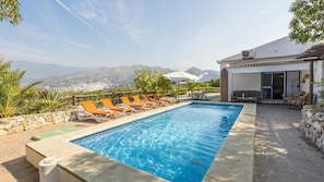 A rectangular swimming pool with clear blue water, surrounded by orange lounge chairs. There is a stone wall on one side and a building with a tiled roof and a door with a metal gate.