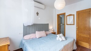 A bedroom with a patterned headboard, a bed with a blue blanket and pink pillows, a bedside table with a lamp, and a wooden door.