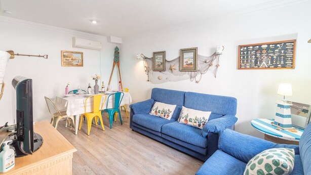 Een woonkamer met een blauwe bank, gele en blauwe stoelen, een eettafel met een witte tafelkleed en een televisie op een houten dressoir.