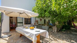 Um pátio com chão de pedra, mesa posta para dois, um guarda-sol branco e um banco.