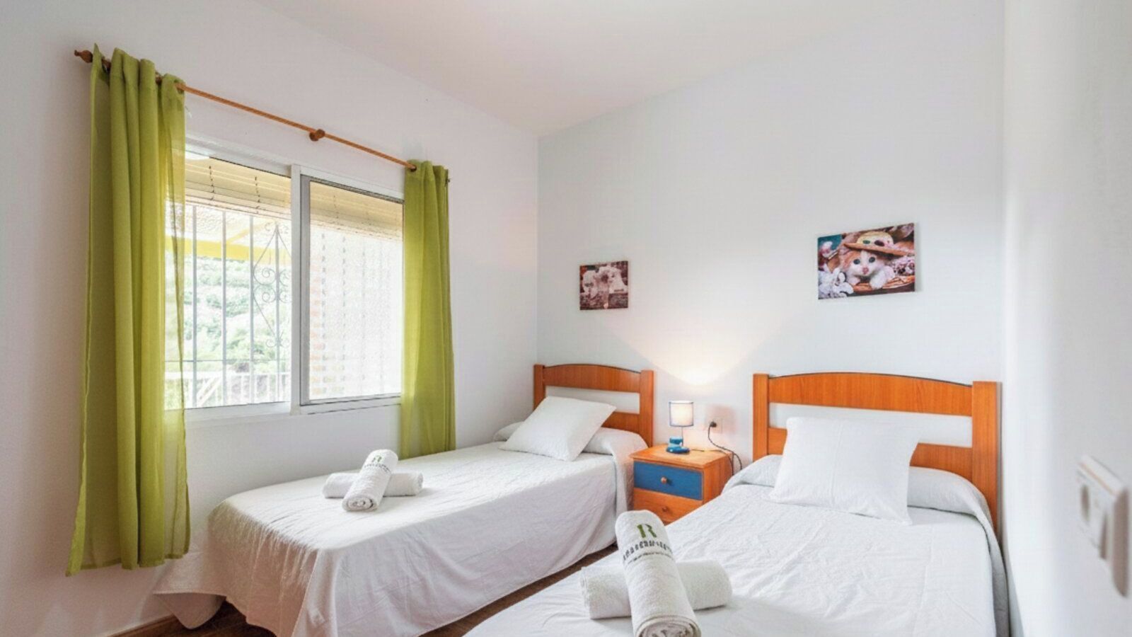 Une chambre d’hôtel avec deux lits simples, une fenêtre avec des rideaux verts, une table de chevet avec une lampe et deux petits tableaux accrochés au mur.