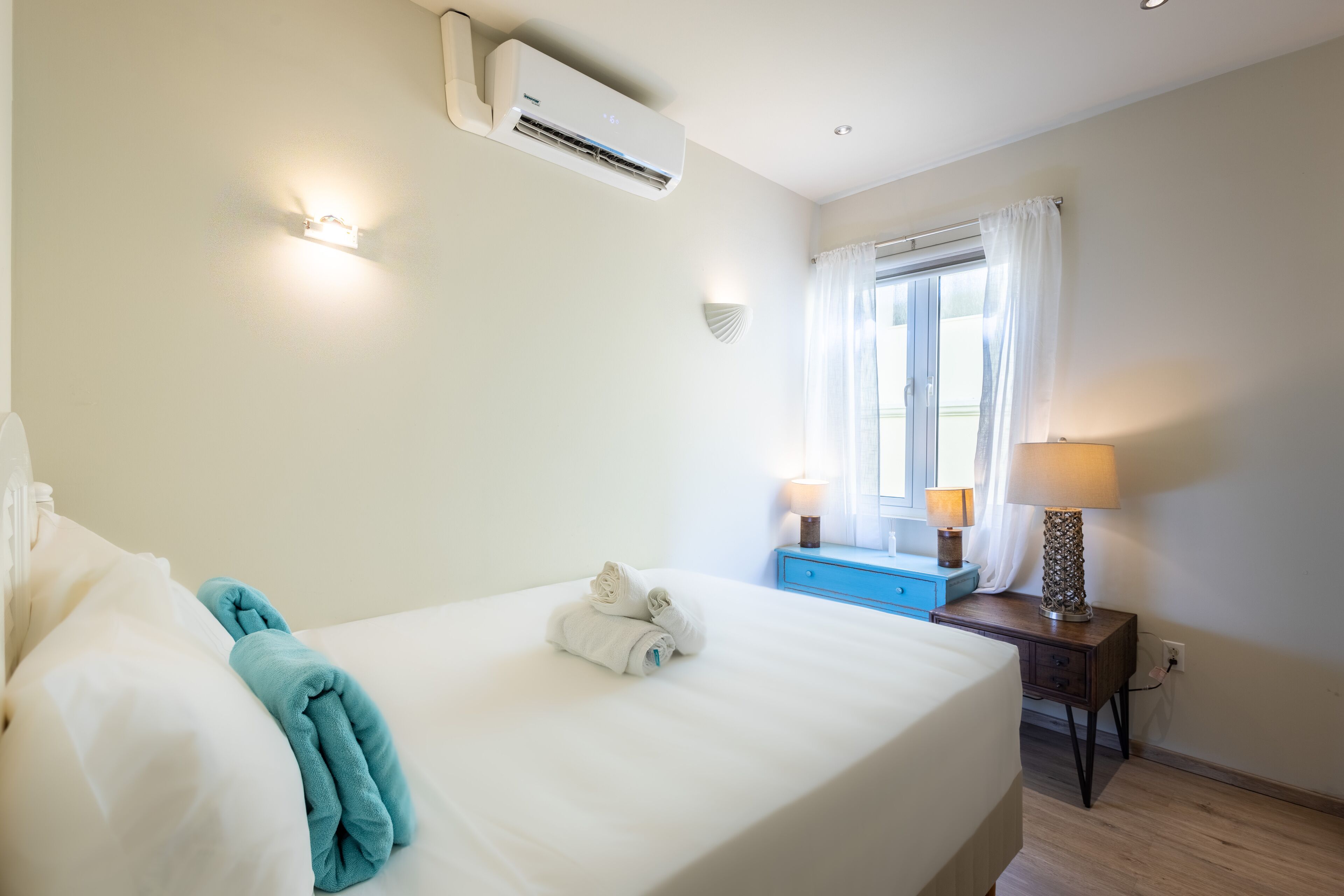 Ein Hotelzimmer mit Bett, Fenster mit Vorhängen, Klimaanlage, Nachttisch mit Lampe und Wandleuchte.