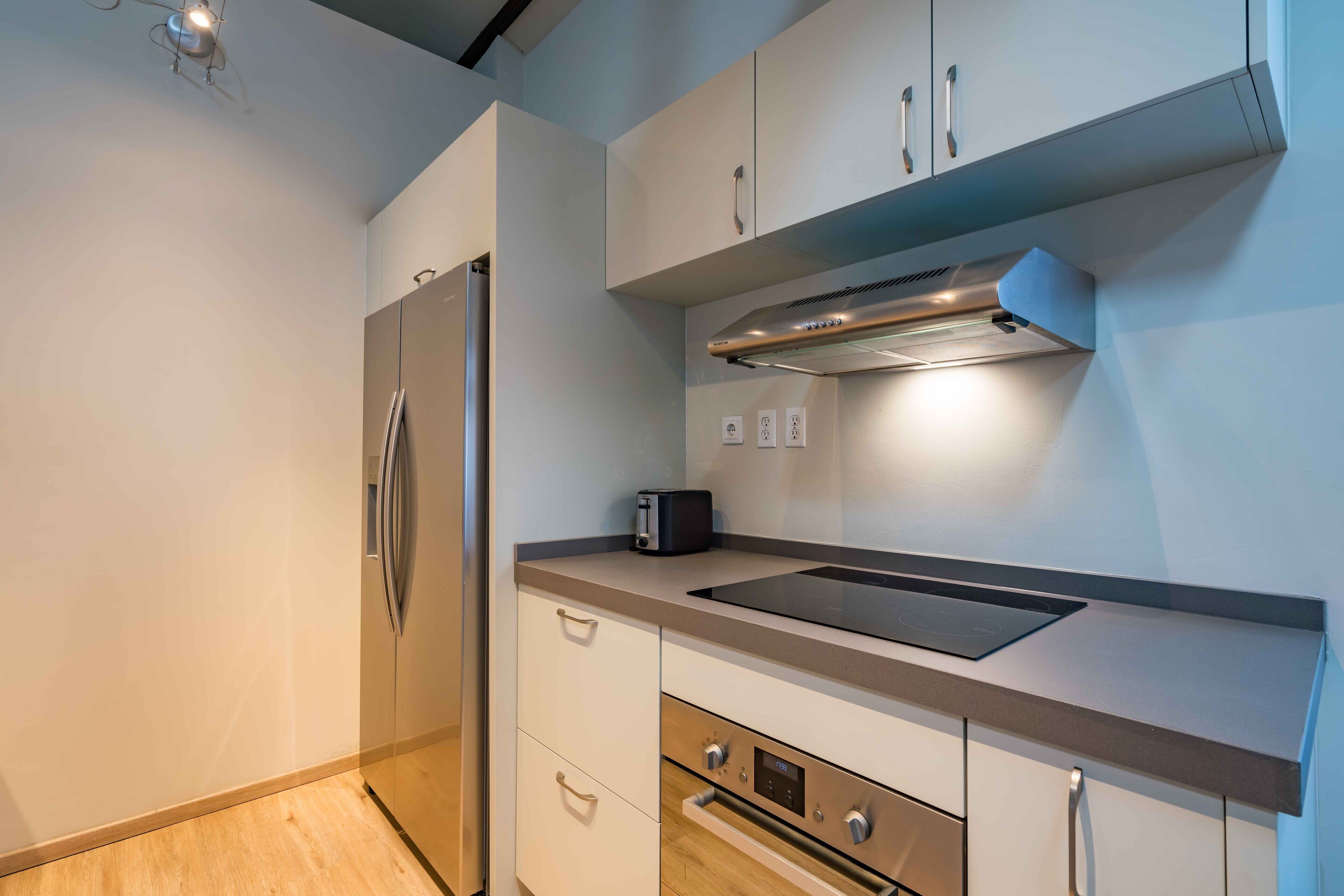 A modern kitchen with a stainless steel refrigerator, a built-in oven, and a microwave. There is a toaster on the countertop, and the cabinets are white with silver handles.