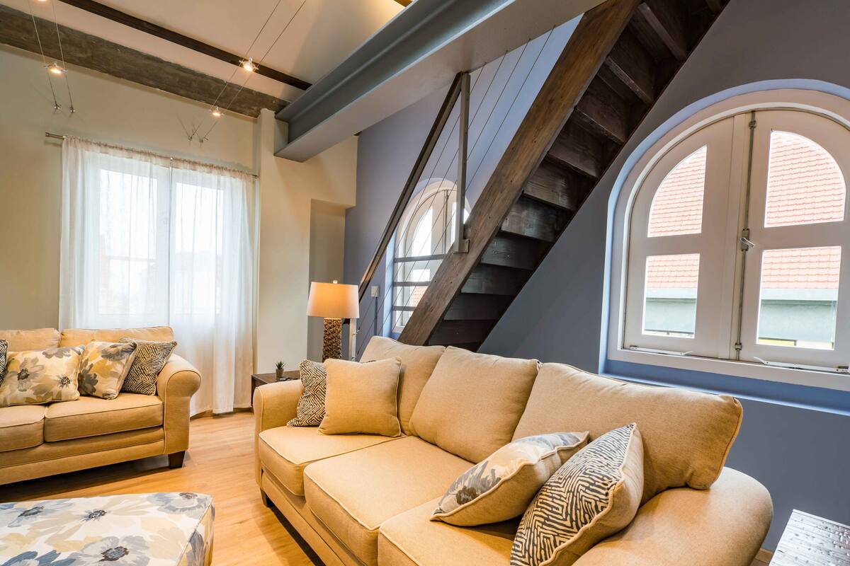 A modern living room with a sofa, a coffee table, and a lamp. There is a window with a view of the outside.