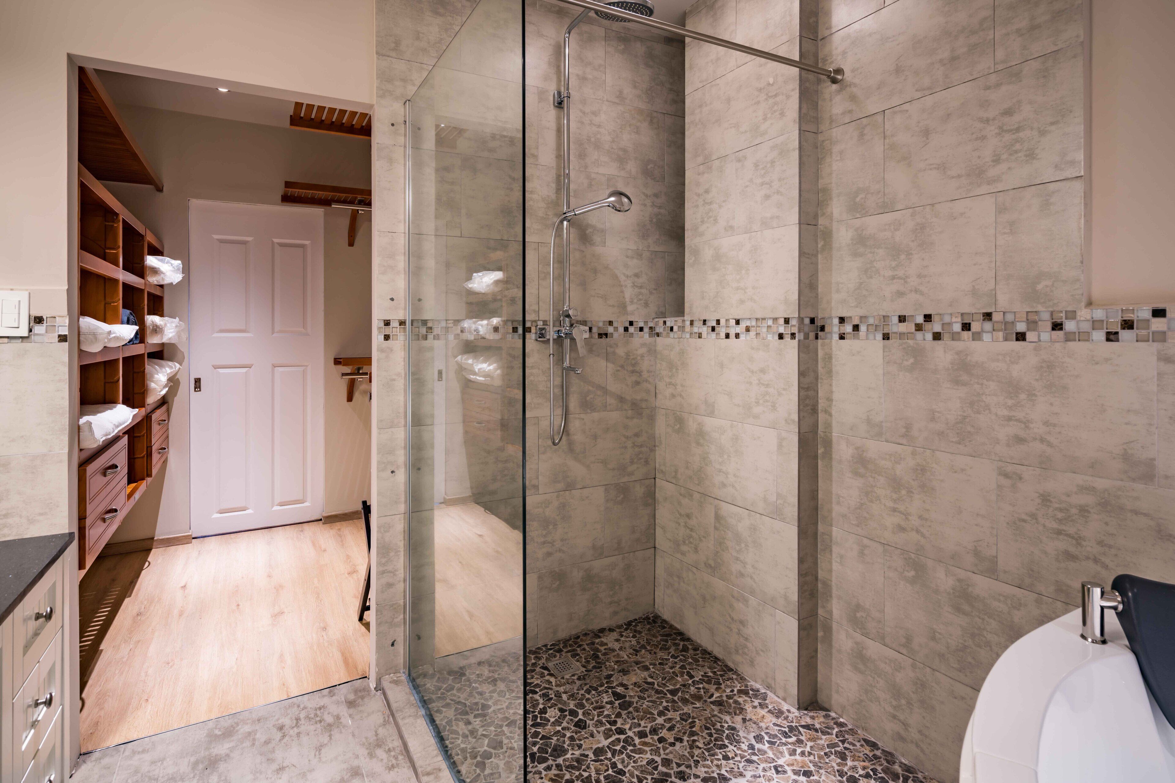 Un baño moderno con cabina de ducha de vidrio, suelo de baldosas y una puerta blanca que da a un espacio de almacenamiento con estanterías.