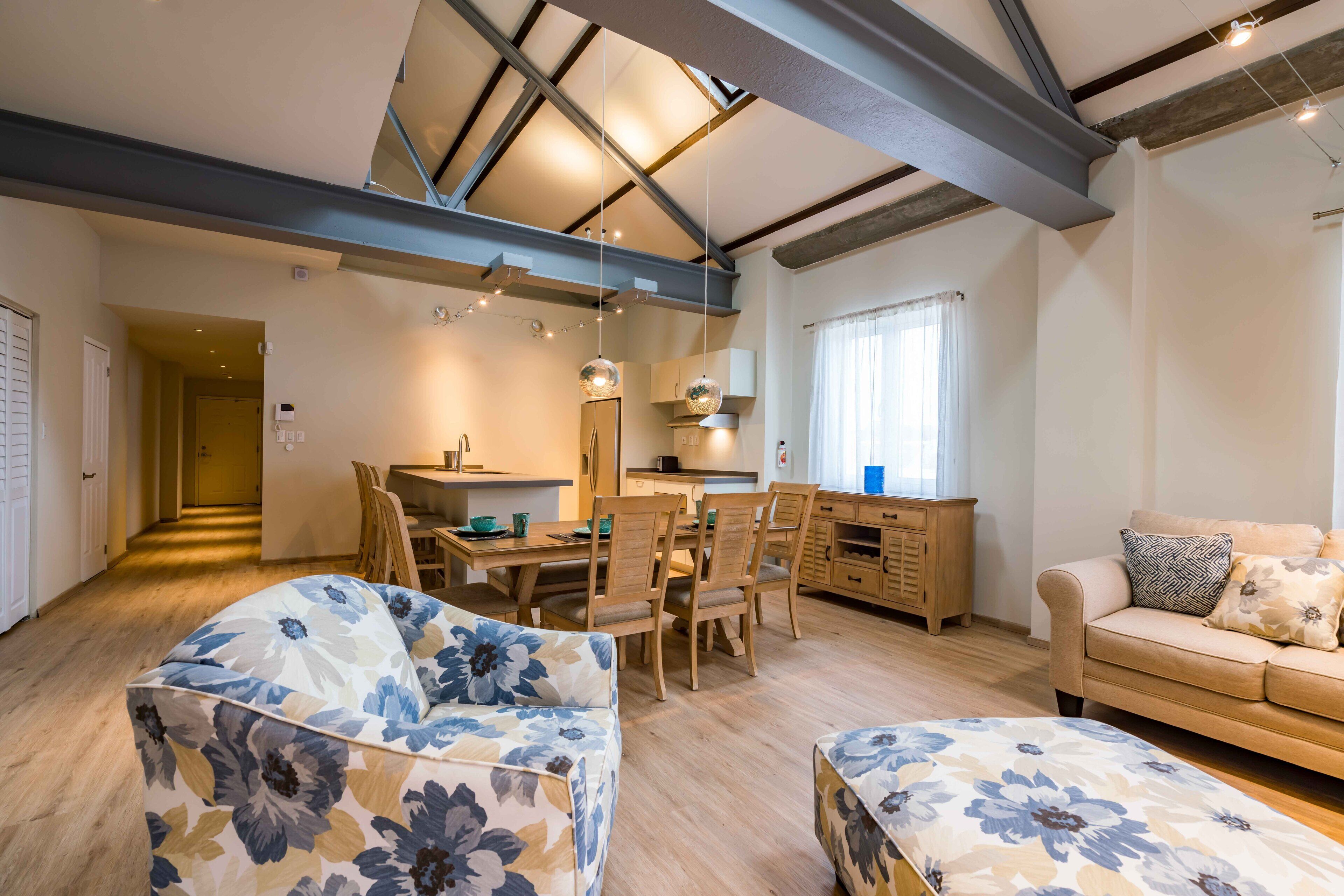 A spacious living area with a high ceiling, wooden dining table, and floral-patterned sofas.