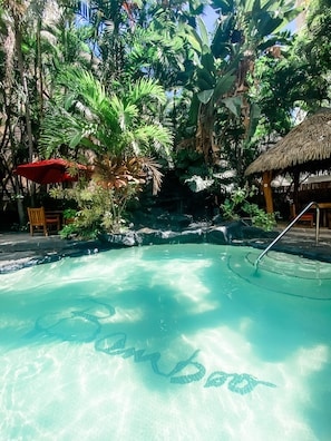 A clear pool surrounded by lush tropical vegetation, a red umbrella, and a thatched-roof structure.