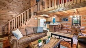 Interior de una cabaña de madera con un sofá de cuero marrón, una mesa de centro de madera y una escalera que lleva a una zona superior.