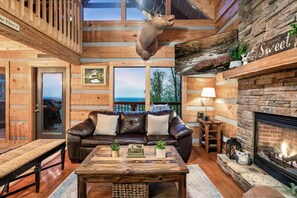 A cozy cabin interior with a leather sofa, a wooden coffee table, a fireplace, and a mounted deer head.