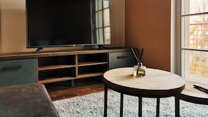 Uma sala de estar moderna com uma grande televisão de tela plana presa a um armário de madeira, uma mesa de madeira redonda com um controle remoto e vista para árvores pela janela.
