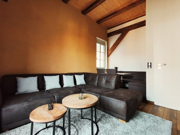 Un salon moderne avec un canapé d’angle, deux petites tables rondes en bois et un escalier dont les poutres en bois sont apparentes.