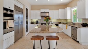 A modern kitchen with white cabinetry, stainless steel appliances, a central island with stools, and a view of greenery outside.