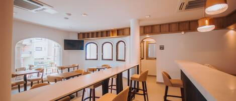A modern dining area with long tables and bar stools, large windows, and a flat-screen TV mounted on the wall.