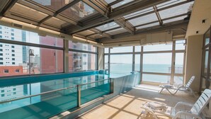 Ein überdachter Pool mit Glasbarriere, sonnendurchflutetem Boden und Liegestühlen.