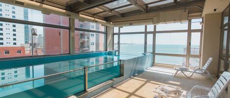 Una piscina interior con barrera de cristal, suelo iluminado por el sol y sillones de descanso.
