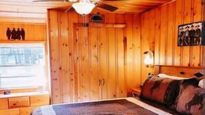 Un dormitorio con paneles de madera, una cama, un ventilador de techo, una mesita de noche y una lámpara de pared.