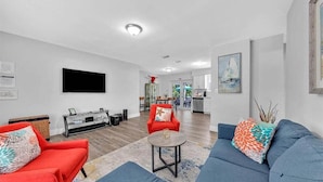 A modern living room with a sofa, armchair, and coffee table. There is a flat-screen TV mounted on the wall, a dining area in the background, and a kitchen visible through an open door. - 3Bed Modern Home Family Friendly Heated Pool (Fort Lauderdale)