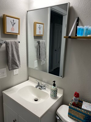 A bathroom with a white sink, a mirror, two towels, a bottle of hand soap, and toiletries on a shelf. - New!! Woodcrest Cottage - Close to Tri-Cities (Kingsport)