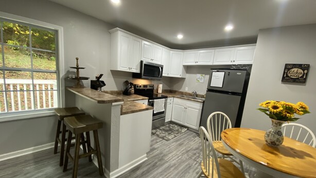 A modern kitchen with white cabinets, a central island, bar stools, a dining table with chairs, a refrigerator, and a microwave. - New!! Woodcrest Cottage - Close to Tri-Cities (Kingsport)