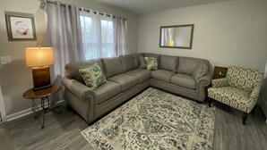 A living room with a sectional sofa, a patterned rug, a side table with a lamp, and a patterned armchair. - New!! Woodcrest Cottage - Close to Tri-Cities (Kingsport)