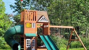 Una casita de juegos de madera con un tobogán y un columpio en un espacio cubierto de césped.