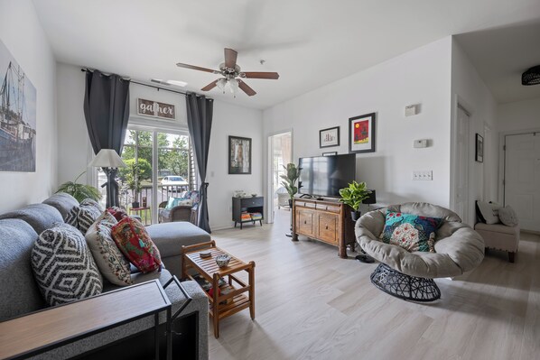 A living room with a sofa, armchair, wooden coffee table, ceiling fan, TV, and a view of the outdoors.