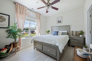 A bedroom with a bed, nightstands, a ceiling fan, a window with curtains, a potted plant, and a small stool.