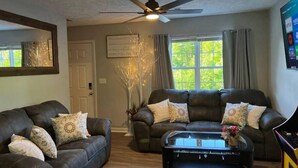 A living room with two dark leather sofas, a glass coffee table, a ceiling fan, a decorative tree, and a window with curtains.