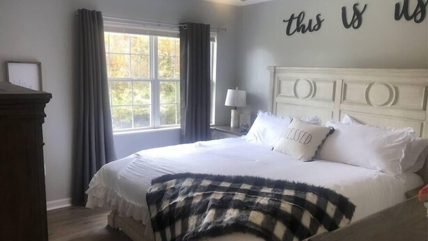 A bedroom with a large bed, a ceiling fan, a window with curtains, and a wall with a motivational quote.