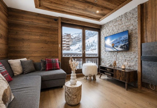 A cozy living room with a wooden ceiling, a stone wall, a large window with a snowy view, a grey sofa with cushions, a round wooden coffee table, and a TV on the wall.
