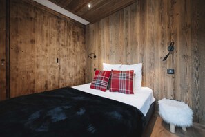 Ein Schlafzimmer mit Holzwand, einem Bett mit weißer Bettwäsche und Karokissen, einer schwarzen Decke, einem weißen, flauschigen Hocker und einer Wandleuchte.