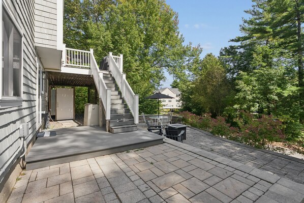 A covered outdoor area with a staircase leading to a deck, surrounded by trees and a patio with outdoor furniture.
