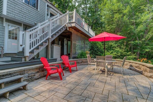 A patio with stone tiles, red Adirondack chairs, a wooden table, and a red umbrella.