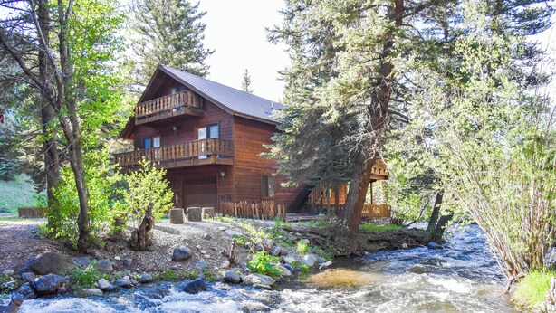 Une cabane en bois dotée d’un balcon, entourée d’arbres et bordée par un ruisseau.