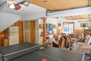 A room with a ping pong table, wooden furniture, a refrigerator, and a window with a view.