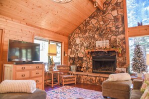 A cozy cabin interior with a stone fireplace, wooden walls, a television, and a fireplace mantel with decorations.