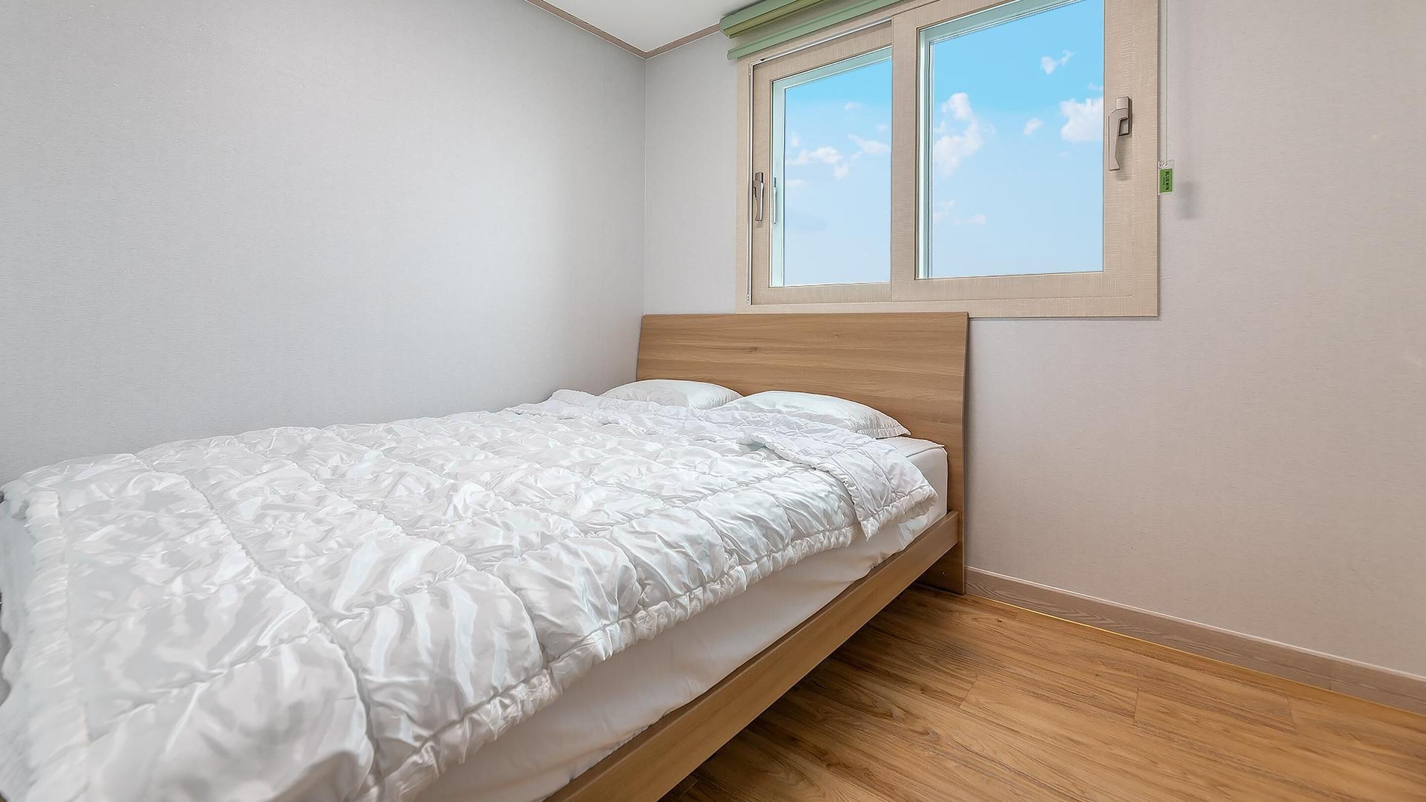 A single bed with a wooden headboard in a room with white walls and a window with a view of the sky.