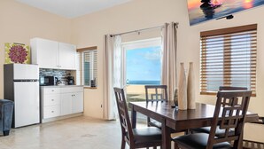 A modern kitchen with white cabinets, a dining table with chairs, and a view of the sea through a large window.