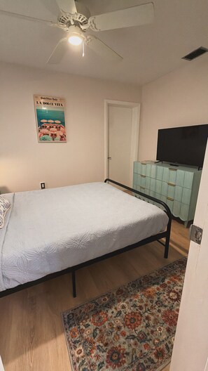 Ein Schlafzimmer mit einem Einzelbett, einem Deckenventilator, einem Fernseher auf einem Schrank und einem Poster an der Wand.