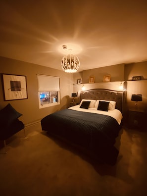 Une chambre à coucher avec un grand lit, un lustre, une fenêtre avec des stores et un tableau encadré au mur.