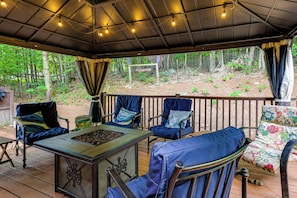 Un patio exterior con terraza de madera, sillas de estructura metálica, una mesa metálica con una bandeja de comida y vistas a una zona boscosa.