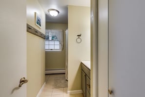 A narrow hallway with a door on the left, a window with a view of a building, and a bathroom with a toilet and sink on the right.