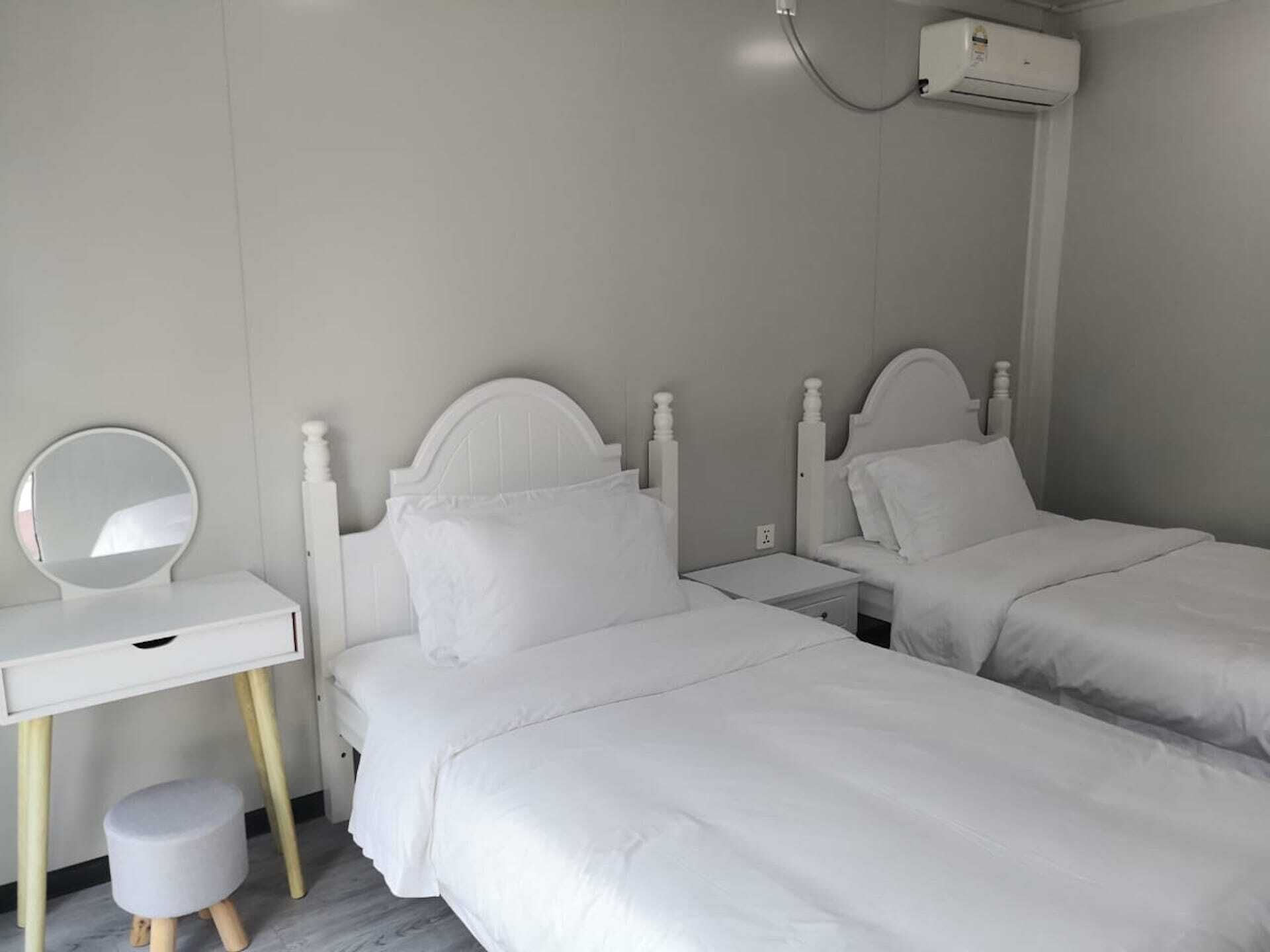Une chambre avec deux lits simples, une table de chevet avec un miroir, un petit tabouret et un climatiseur fixé au mur.