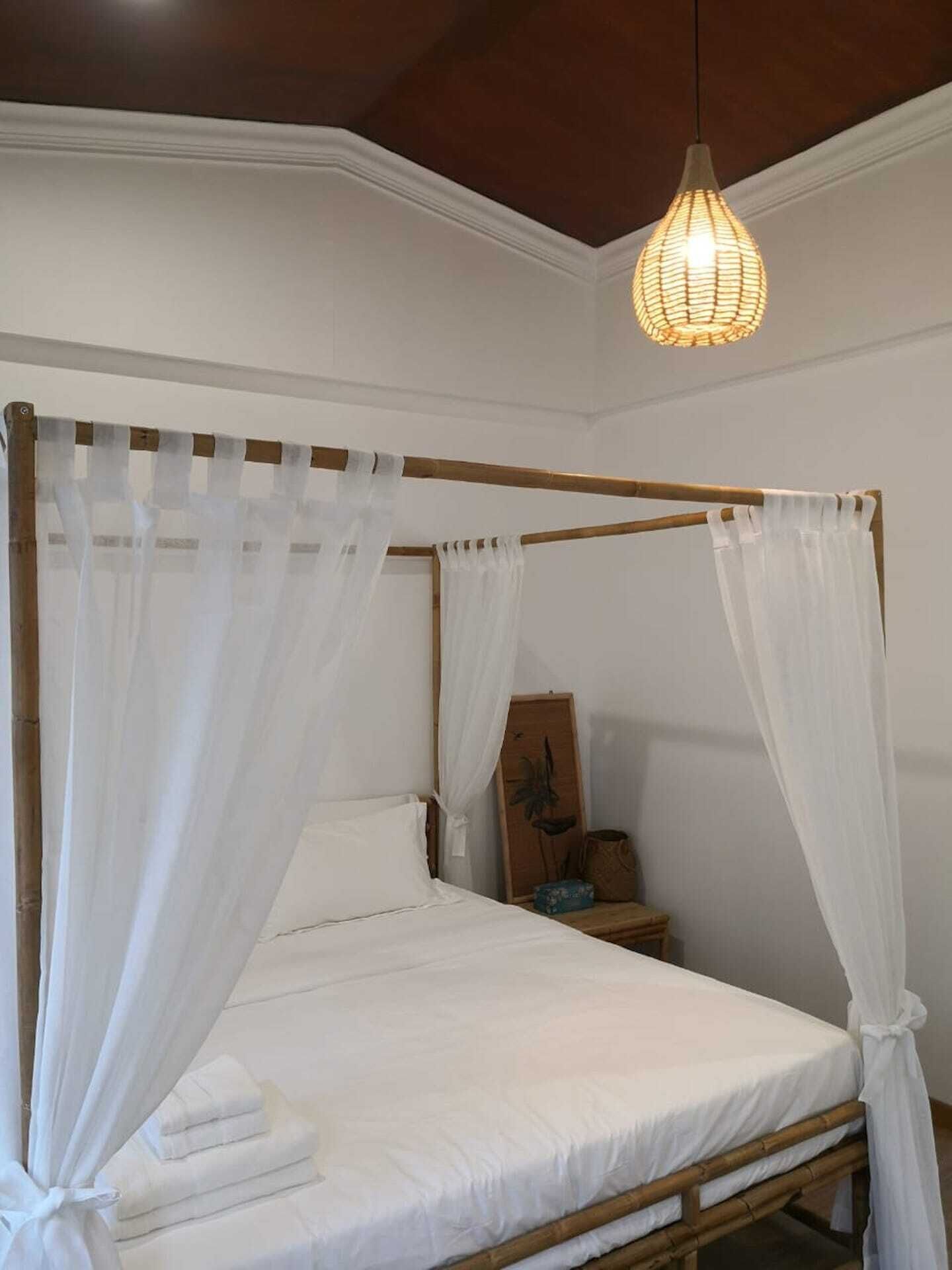 Un lit à baldaquin avec du linge de lit blanc et des rideaux vaporeux, une table de chevet en bois avec une lampe, et un luminaire suspendu.